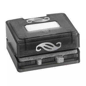 May include: A dark gray, rectangular paper punch with a silver decorative design. The punch has a clear top with a rectangular opening and a silver design. The bottom section is a silver metal base.