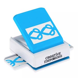 May include: A blue and white paper punch with a decorative design. The punch has a blue top with a white design and the words "creative memories" printed on the side. A piece of paper with the punched design is visible.