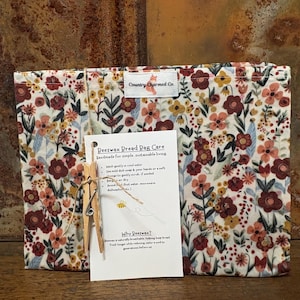 May include: A floral patterned bread bag with a tag that reads "Beeswax Bread Bag Care" and "Country Charmed Co." The bag is held closed with a wooden clothespin. The floral pattern includes red, yellow, and white flowers.