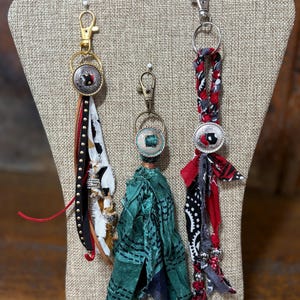 May include: Three keychains with decorative fabric tassels and metal accents. The keychains feature a variety of colors, including red, black, white, and green. Each keychain has a metal clasp for easy attachment.