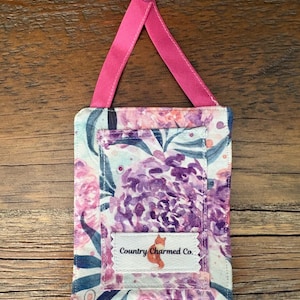 May include: A rectangular floral fabric item with a pink ribbon and button for hanging. The fabric features a watercolor design with purple and pink flowers, and the text "Country Charmed Co." is visible on a small label.