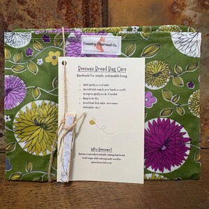 May include: A green beeswax bread bag with a floral pattern in yellow, purple, and white. The bag is accompanied by a care instruction card held by a wooden clip. The card includes text about washing and care instructions.