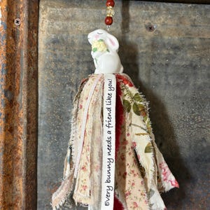 May include: A white ceramic bunny figurine holding flowers, suspended by a red cord with wooden beads. The bunny is attached to a fabric tassel with floral patterns and the text "Every bunny needs a friend like you!"