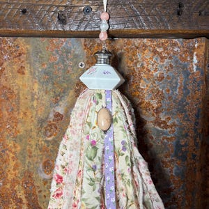 May include: A handmade tassel ornament with a floral fabric design in pink, green, and cream. The ornament features a white ceramic top with pink floral accents, a gemstone pendant, and pink and clear beads. A white ribbon is used for hanging.