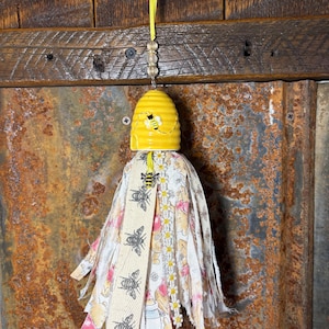 May include: A decorative tassel featuring a yellow ceramic beehive with a painted bee design. The tassel is made of fabric strips in cream, pink, and white, adorned with bee and daisy patterns. A small bee charm is attached.