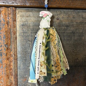 May include: A decorative tassel featuring a white ceramic bunny holding a pink flower. The tassel is made of fabric strips in various colors, including blue, green, and yellow, with a cream-colored ribbon that reads "Somebunny Loves You."