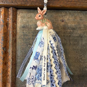 May include: A decorative tassel featuring a ceramic bunny figurine atop fabric strips in shades of blue and white. The tassel includes ribbons, lace, and a tag that reads "What's up, hop stuff?" A blue cord with beads is attached.