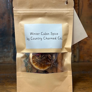 Puede incluir: Una bolsa de papel marrón con una ventana transparente que muestra especias secas. Las especias incluyen una rodaja de naranja seca, anís estrellado y palitos de canela. La etiqueta blanca dice "Winter Cabin Spice by Country Charmed Co."