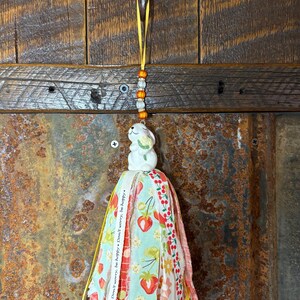 May include: A decorative tassel with a ceramic bunny figurine on top. The tassel features fabric strips with strawberry and cherry patterns, and the text "Don't worry, be happy." A yellow ribbon and beaded strand are attached.