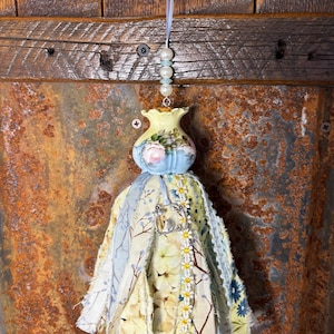 May include: A handmade tassel ornament with a small, light blue and yellow vase-shaped top adorned with floral designs. The tassel features various fabrics in shades of yellow, blue, and white, with a small bird charm and daisy trim. A string of beads and ribbon are attached.