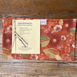 May include: A beeswax bread bag with a floral pattern in shades of red, pink, and green. A care instruction card is attached with a wooden clothespin. The bag is designed for simple, sustainable living.