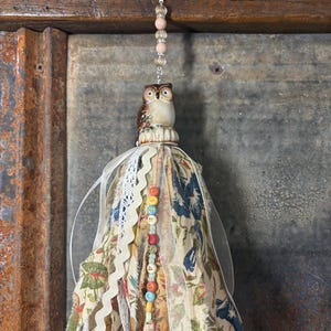 May include: A decorative tassel featuring a ceramic owl figurine, floral fabric, lace, and colorful beads. The tassel is suspended by a cream-colored ribbon and is designed for hanging. The owl has brown and cream coloring.