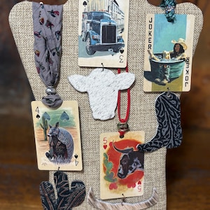May include: A display of handmade Western-themed jewelry. The collection includes playing card pendants with images of a truck, a woman in a tub, and an armadillo. Also featured are cowhide and leather accents, including a cow head, a bull head, and a cactus.