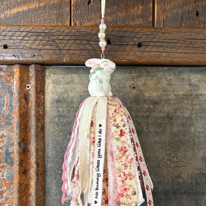 May include: A decorative Easter-themed tassel featuring a white ceramic bunny holding flowers. The tassel includes pink and floral fabric ribbons, lace, and a ribbon with the text "No bunny likes you like I do". The tassel is suspended by a cream-colored cord with beads.