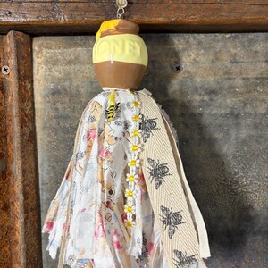 May include: A decorative honey pot tassel with a yellow and brown ceramic pot, the word "HONEY" is visible. The tassel features fabric with a Winnie the Pooh design, a daisy chain, and a beige ribbon with black bee graphics. A yellow beaded string is attached.