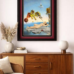 Puede incluir: Impresión enmarcada de una pintura surrealista con una figura desnuda sobre una roca, con tigres y otros elementos flotando en el cielo. La obra de arte está enmarcada en negro con un passe-partout burdeos, expuesta en un mueble de madera.