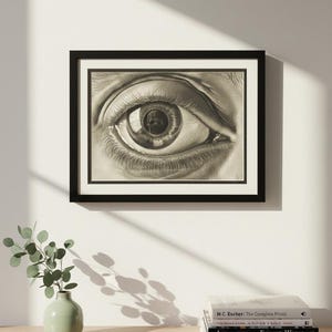 Puede incluir: Una impresión en blanco y negro de un ojo humano enmarcada en negro. La obra de arte está colgada en una pared blanca. Un pequeño jarrón verde con vegetación y una pila de libros están sobre una superficie de madera debajo.