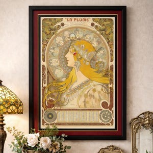 Puede incluir: Impresión artística enmarcada de estilo Art Nouveau. La obra presenta a una mujer con largo cabello rubio y un tocado decorativo. La impresión está en un marco negro con un borde interior rojo. El texto "LA PLUME" está en la parte superior.