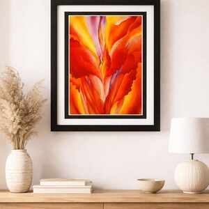 Puede incluir: Impresión artística floral abstracta en un marco negro. La obra presenta tonos vibrantes de rojo, naranja y amarillo, con toques de morado. La pintura se exhibe en una pared blanca sobre una mesa de madera.