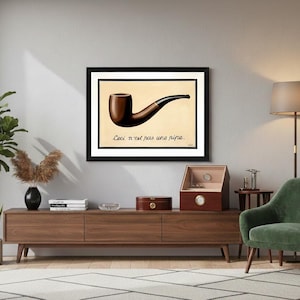 May include: A framed print featuring a brown pipe against a beige background, with the French text "Ceci n'est pas une pipe" below. The artwork is displayed above a wooden cabinet.
