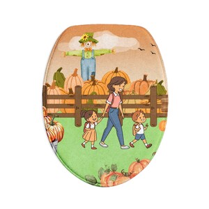 Op de afbeelding: Een ovaalvormige badmat met een kleurrijke illustratie van een vogelverschrikker in een pompoenveld met een houten hek, een ouder en twee kinderen die samen wandelen. De scène bevat oranje pompoenen, groen gras en een zachte zonsondergang.