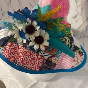 May include: A patchwork sun hat with a wide brim, decorated with blue and white flowers, and various colourful fabric patterns. The hat features a teal band and a blue trim. The crown is pink with white polka dots.