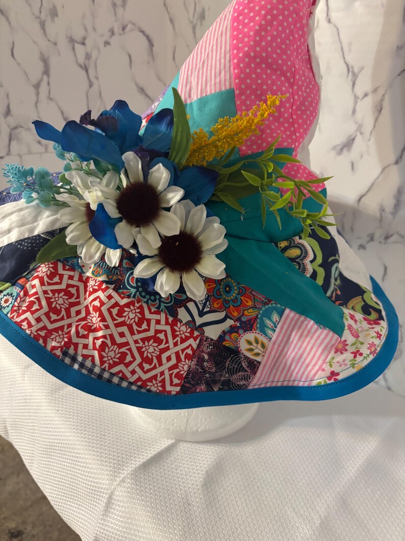 May include: A patchwork sun hat with a wide brim, decorated with blue and white flowers, and various colourful fabric patterns. The hat features a teal band and a blue trim. The crown is pink with white polka dots.