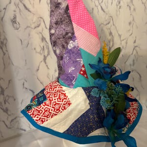 May include: A patchwork bonnet with a variety of fabrics in purple, pink, teal, red, and white. The bonnet is decorated with blue flowers and greenery. The brim is trimmed with a blue border. The bonnet is displayed on a white styrofoam head.