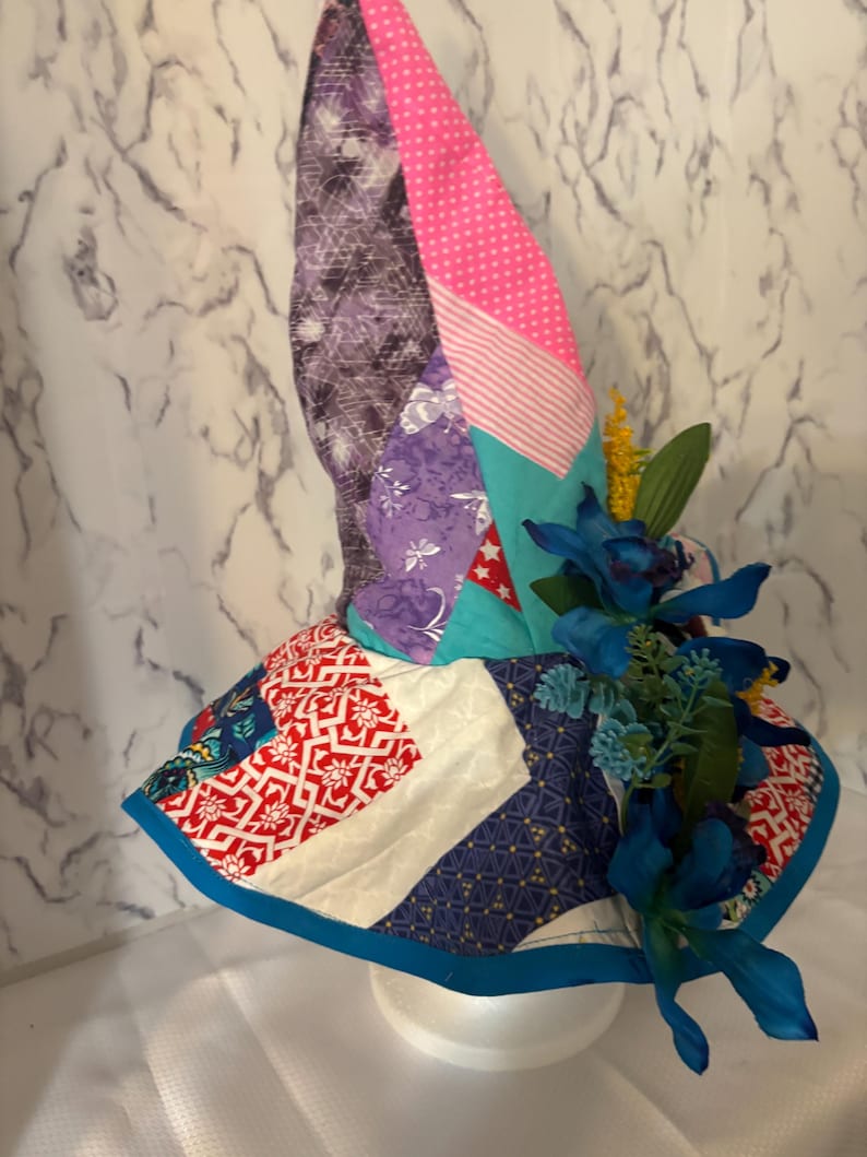 May include: A patchwork bonnet featuring fabrics in purple, pink, teal, red, and white. The bonnet is adorned with blue flowers and greenery. The brim has a blue border. The bonnet is displayed on a white polystyrene head.