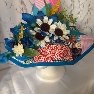 May include: A decorative witch hat with a patchwork design in pink, red, teal, and patterned fabrics. The hat is adorned with blue and white flowers, yellow sprigs, and green leaves. The brim is trimmed with a bright blue border.