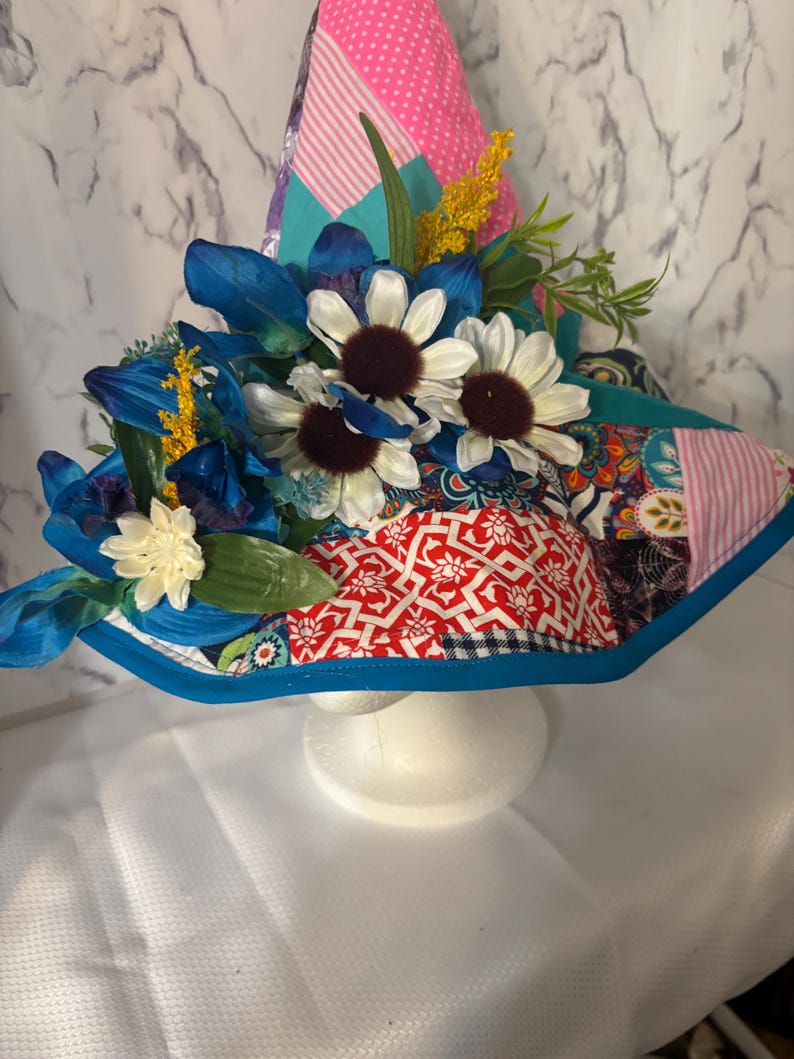 May include: A decorative witch hat with a patchwork design in pink, red, teal, and patterned fabrics. The hat is adorned with blue and white flowers, yellow sprigs, and green leaves. The brim is trimmed with a bright blue border.