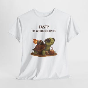 Può includere: T-shirt bianca con una tartaruga dei cartoni animati con occhiali da sole e un jetpack, con la scritta "FAST? I'M WORKING ON IT." La tartaruga è verde e marrone, con una mela accanto. Il jetpack ha una fiamma che esce.