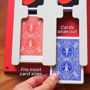 Op de afbeelding: Een rode plastic speelkaarthouder met witte verdelers en sleuven voor kaartspellen. De houder toont de tekst "Cards slide out" en "Fits most card sizes" in het wit. Een blauwe speelkaart wordt uit een van de sleuven getrokken.