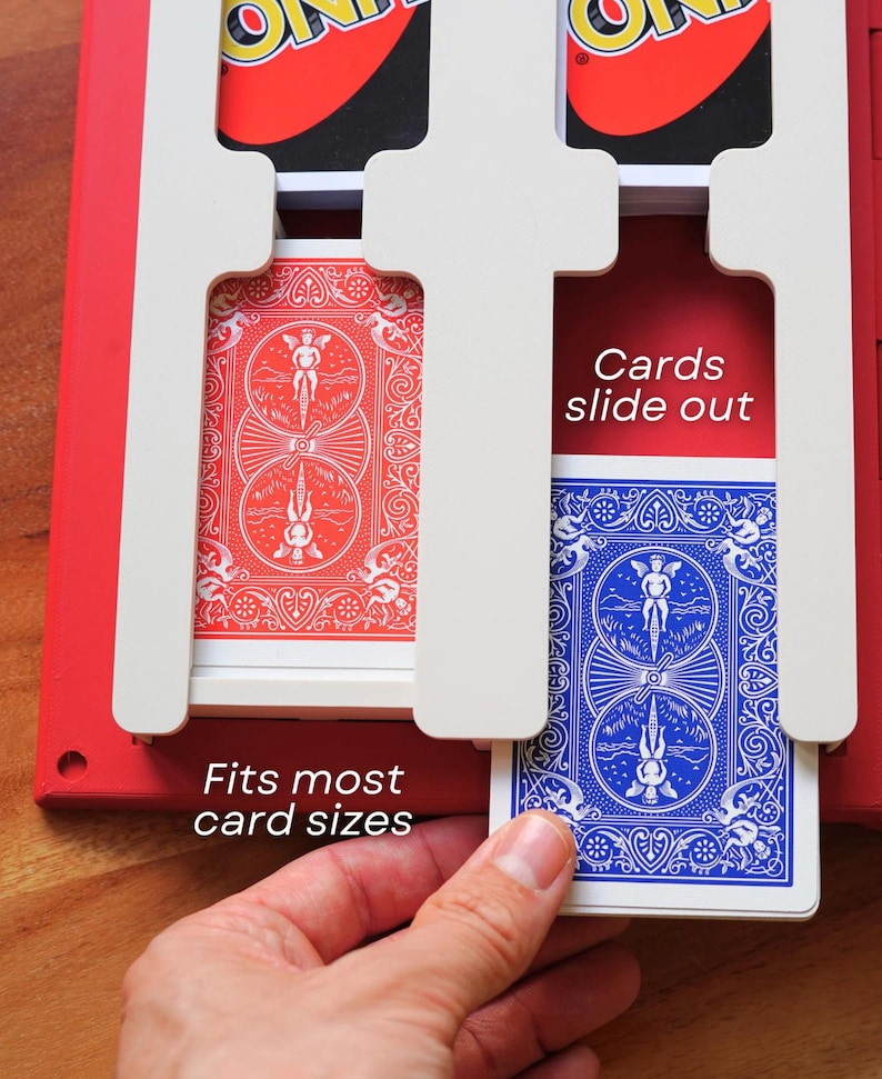 Op de afbeelding: Een rode plastic speelkaarthouder met witte verdelers en sleuven voor kaartspellen. De houder toont de tekst "Cards slide out" en "Fits most card sizes" in het wit. Een blauwe speelkaart wordt uit een van de sleuven getrokken.