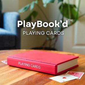 Op de afbeelding: Een rode rechthoekige doos ontworpen als een boek met de tekst PLAYING CARDS op de rug gedrukt. De doos rust op een houten oppervlak met twee speelkaarten zichtbaar, &eacute;&eacute;n met een rood harten symbool en de andere met een rood patroon.