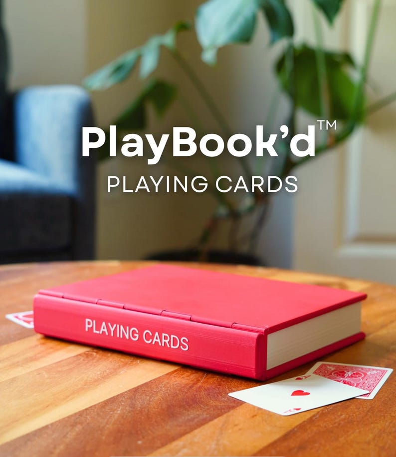 Op de afbeelding: Een rode rechthoekige doos ontworpen als een boek met de tekst PLAYING CARDS op de rug gedrukt. De doos rust op een houten oppervlak met twee speelkaarten zichtbaar, &eacute;&eacute;n met een rood harten symbool en de andere met een rood patroon.