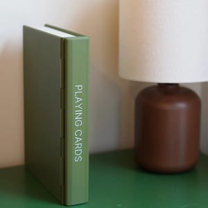 Op de afbeelding: Een rechthoekige olijfgroene opbergdoos voor speelkaarten, rechtopstaand op de zijkant. Op de rug van de doos staat de tekst PLAYING CARDS in witte hoofdletters gedrukt. De doos heeft een gladde, effen gekleurde buitenkant.