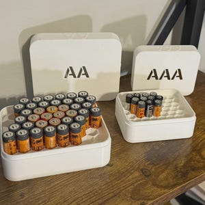May include: White battery organizers with AA and AAA labels. The organizers hold multiple batteries, with the AA organizer holding a larger quantity. The batteries are orange and black.