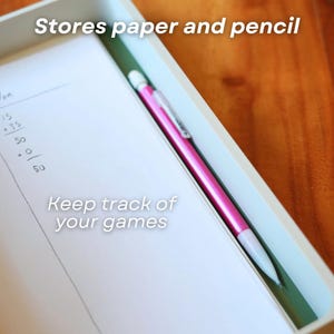 Op de afbeelding: Een wit rechthoekig dienblad met een stapel wit papier met handgeschreven spelscores en een felroze vulpotlood. Op het dienblad staat de tekst Stores paper and pencil en Keep track of your games gedrukt.