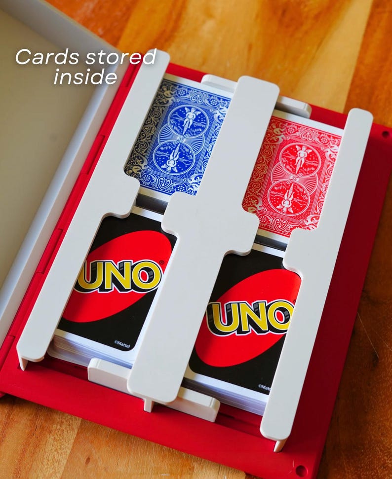 Op de afbeelding: Een rode plastic opbergdoos voor speelkaarten met een witte verdeler. De doos bevat twee stapels UNO-kaarten en twee stapels speelkaarten met blauwe en rode patronen. De tekst "Cards stored inside" is bovenaan in de afbeelding zichtbaar.