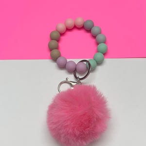 May include: A pastel-colored beaded bracelet with a fluffy pink pom-pom keychain. The bracelet features a gradient of colors, including pink, gray, green, and purple. The pom-pom is a vibrant pink color and is attached to the bracelet with a silver clasp.