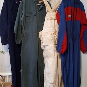 May include: Selection of workwear and outerwear hanging on a rail. Includes a navy blue jacket, a green jumpsuit, off-white overalls, and a blue and red ski suit. The garments are displayed on hangers against a plain backdrop.