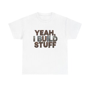 May include: White t-shirt with the text "YEAH, I BUILD STUFF" in a distressed, industrial-style font. The text is in shades of brown and gray, giving it a weathered appearance. The shirt is a classic crew neck style.