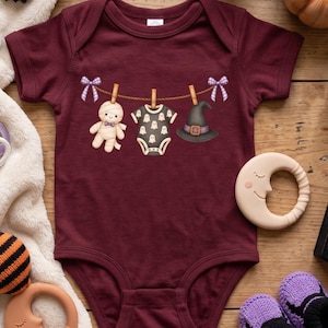 May include: A maroon short-sleeved baby bodysuit featuring a graphic print of a clothesline with a mummy doll, a small ghost-patterned bodysuit, and a witch hat hanging from wooden clothespins, accented by two purple and white checkered bows.