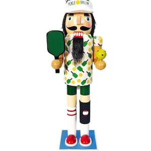 May include: A wooden nutcracker figurine dressed in a pickleball-themed outfit. The nutcracker wears a white visor with "PICKLE BALLIN" printed on it, a patterned shirt, green shorts, and holds a paddle and ball.