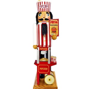 May include: A wooden popcorn vendor nutcracker with a red and white striped uniform and a popcorn-shaped hat. The nutcracker holds a sign that says "Fresh Popcorn". The base of the nutcracker is a red popcorn cart with yellow wheels.