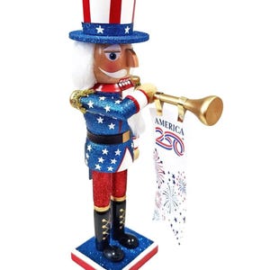 May include: A patriotic Uncle Sam nutcracker figurine. The figurine wears a blue jacket with white stars, red pants, and a tall red, white, and blue hat. It holds a golden trumpet and a banner that reads "America 250".