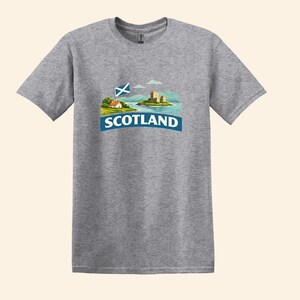 May include: A heather gray t-shirt featuring a graphic of a Scottish landscape with a castle, a flag, and the word "SCOTLAND" in a blue banner. The design is centered on the front of the short-sleeved shirt.