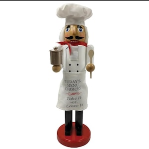 May include: A decorative chef nutcracker figurine. The figurine wears a white chef's hat and uniform, a red scarf, and a white apron with the text "Today's Menu Choices - Take It - or - Leave It". It holds a brown spice grinder and a wooden spoon.