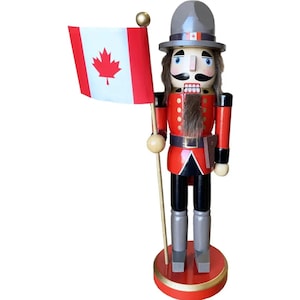 May include: A wooden nutcracker figurine in a red and black uniform, holding a Canadian flag. The figurine has a gray hat, a long mustache, and a faux fur beard. It stands on a red base with gold trim, a festive and patriotic decoration.
